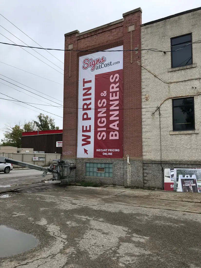 Tall vertical vinyl banner reading “We print signs & banners” on a brick building